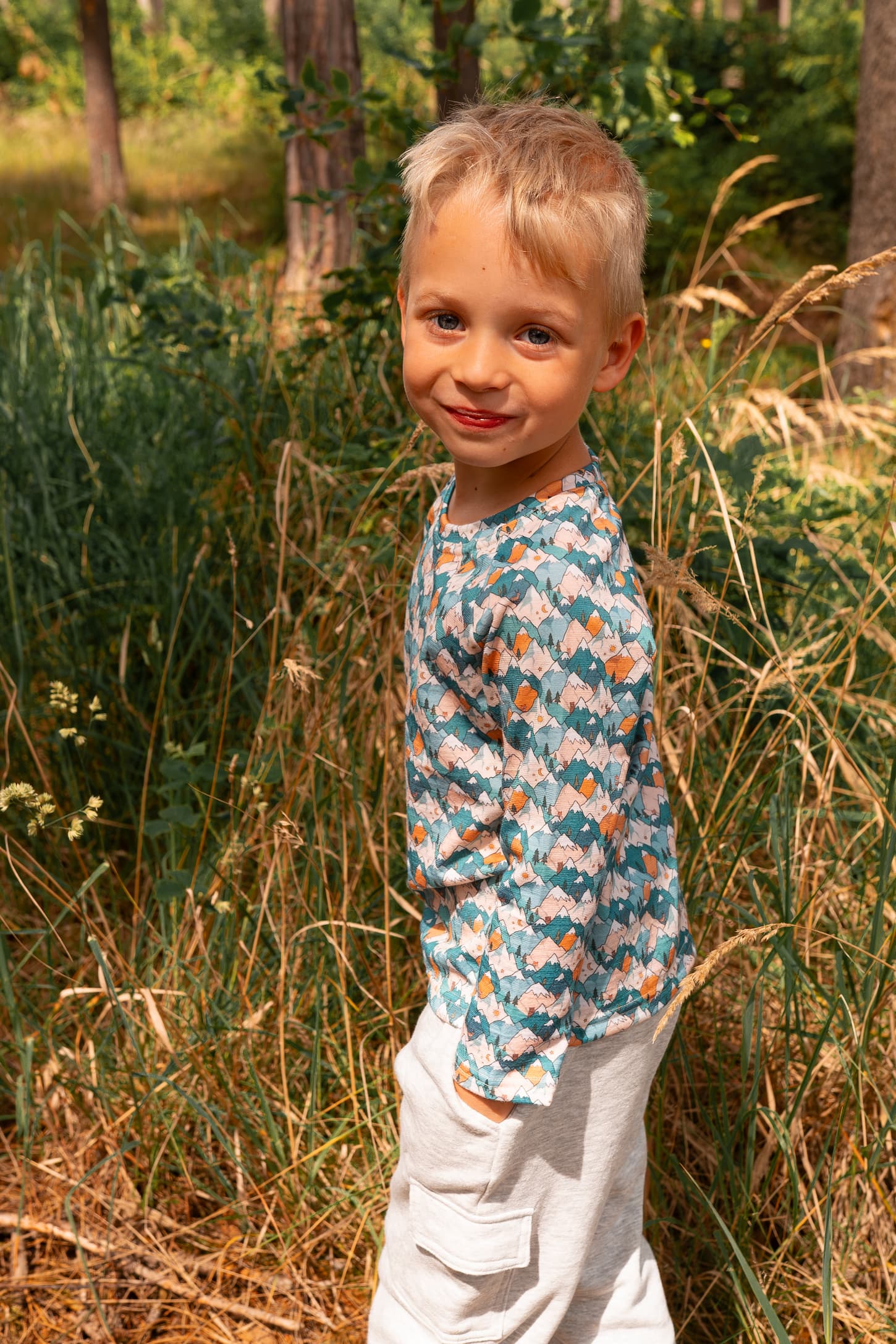 Junge trägt ein Merino Kinder-Langarmshirt mit blauem Bergmotiv von Bellibell im Wald.