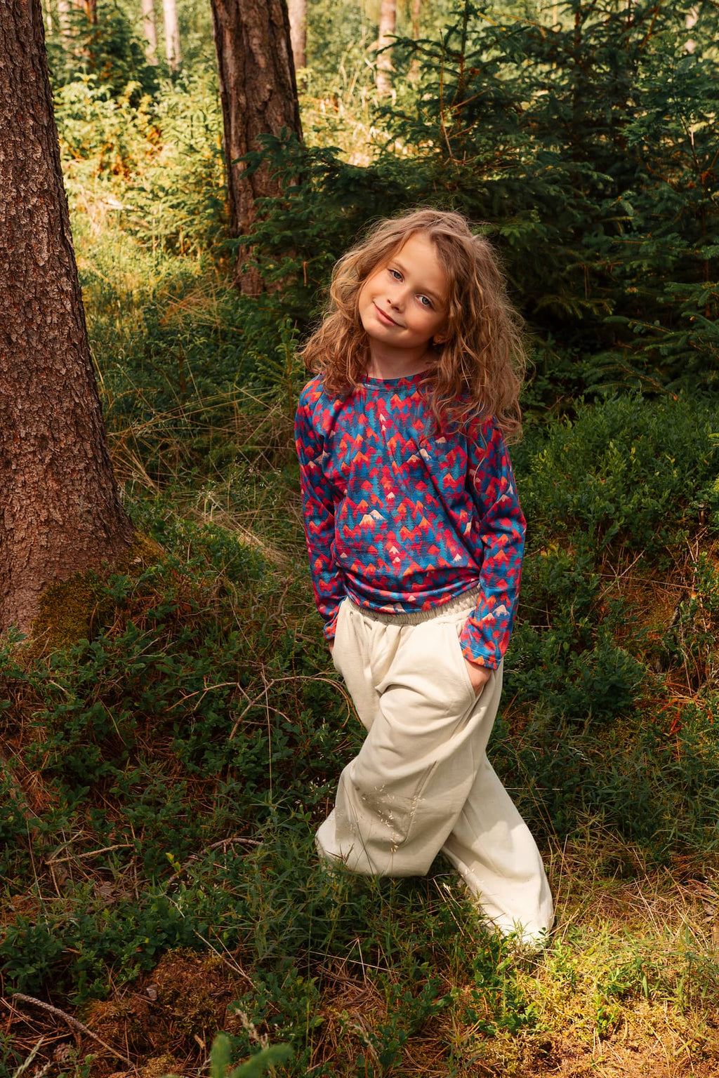 Mädchen trägt ein Merino Kinder-Langarmshirt mit buntem Bergmotiv von Bellibell und steht im Wald.