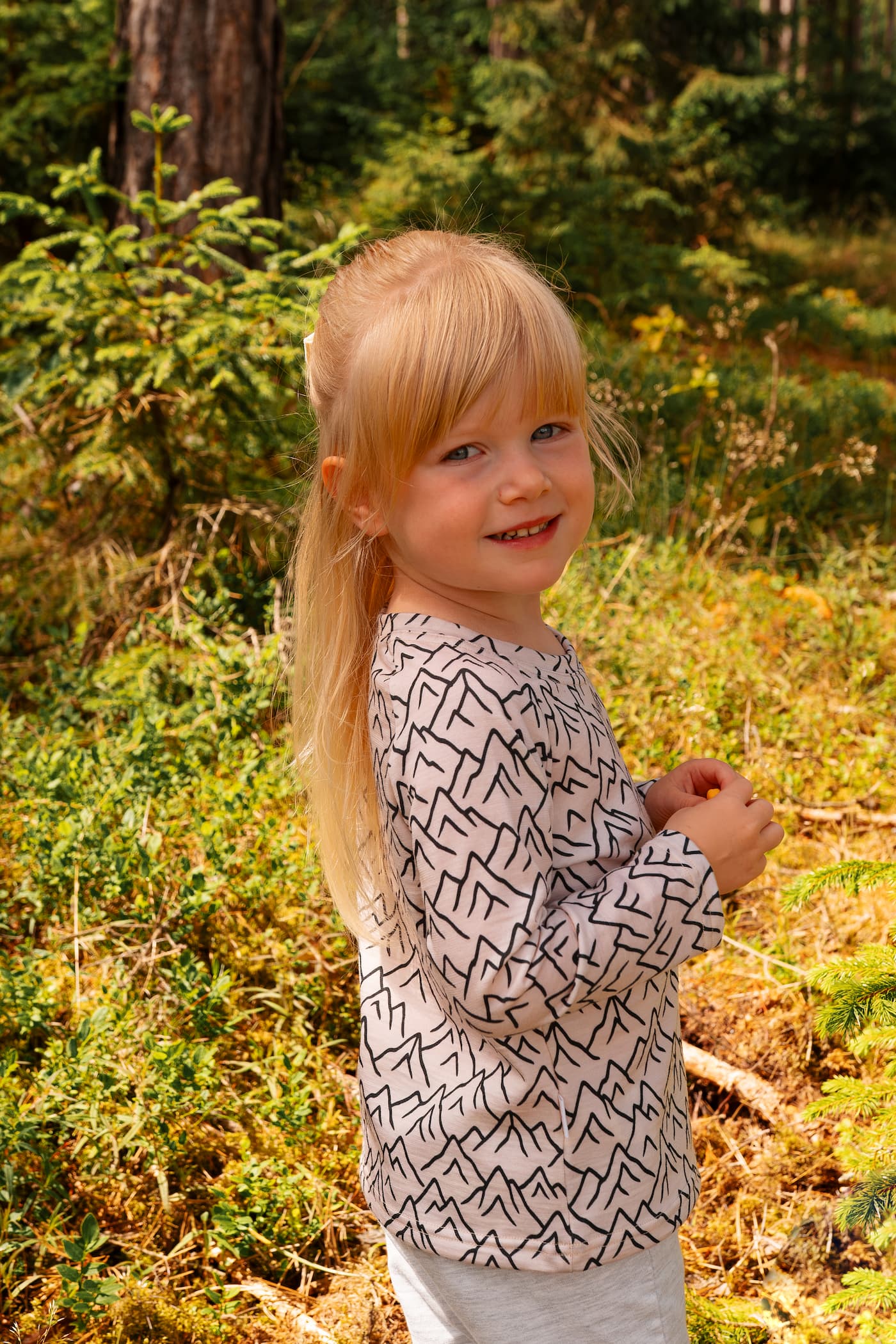 Mädchen trägt ein Merino Kinder-Langarmshirt mit grauem Bergmotiv von Bellibell im Wald.