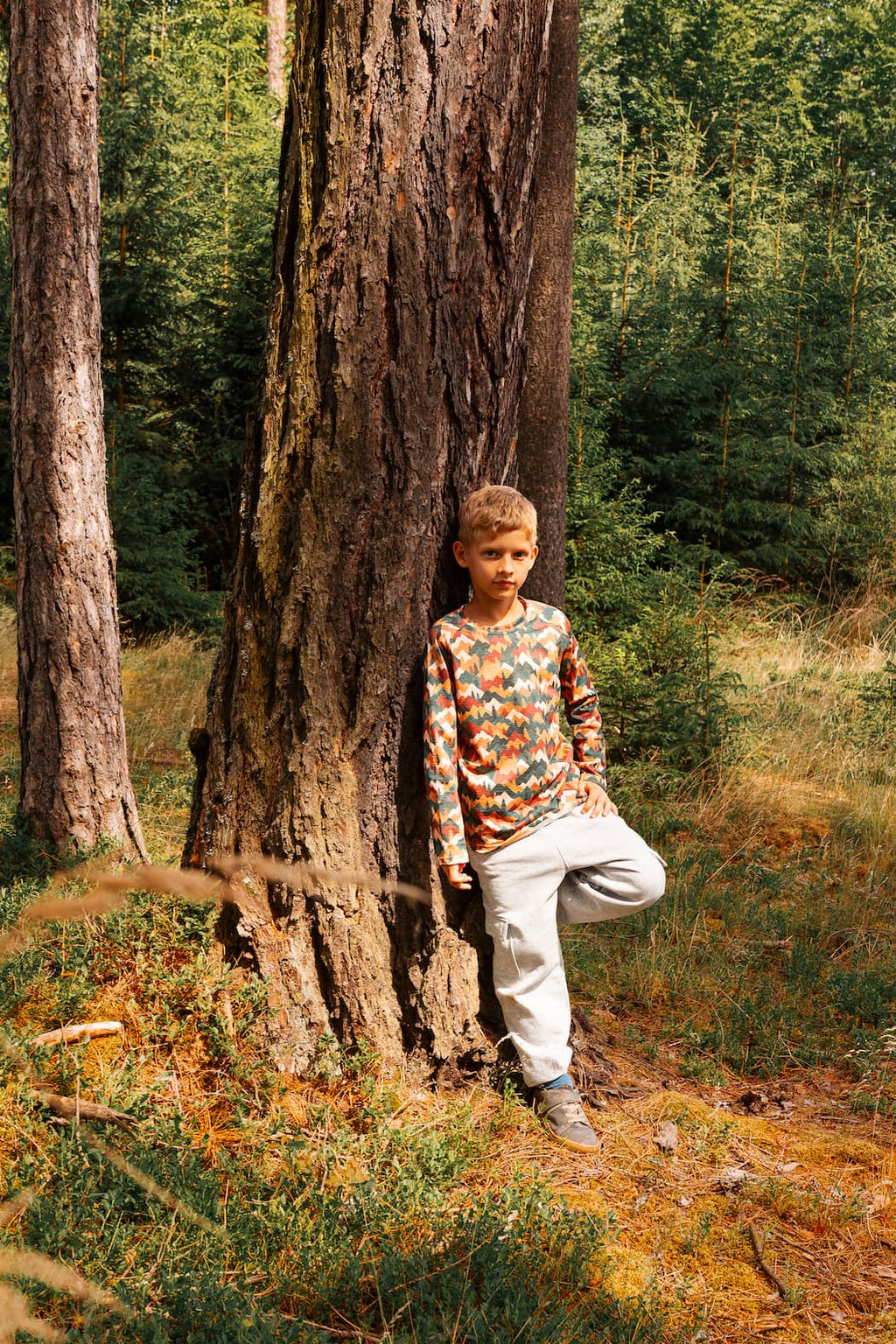 Junge trägt ein Merino Kinder-Langarmshirt mit Herbst-Bergmotiv von Bellibell und lehnt an einem Baum im Wald.