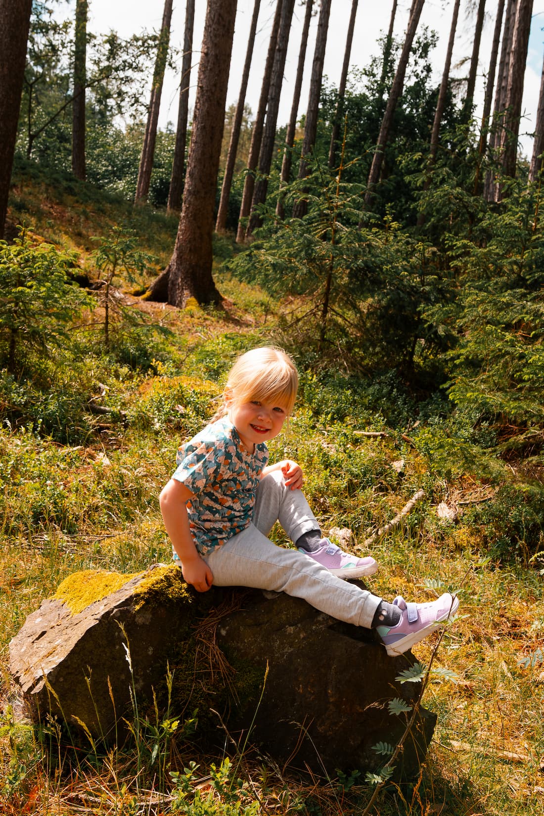 Mädchen trägt ein Merino Kinder T-Shirt mit blauem Bergmotiv von Bellibell und sitzt auf einem Felsen im Wald.
