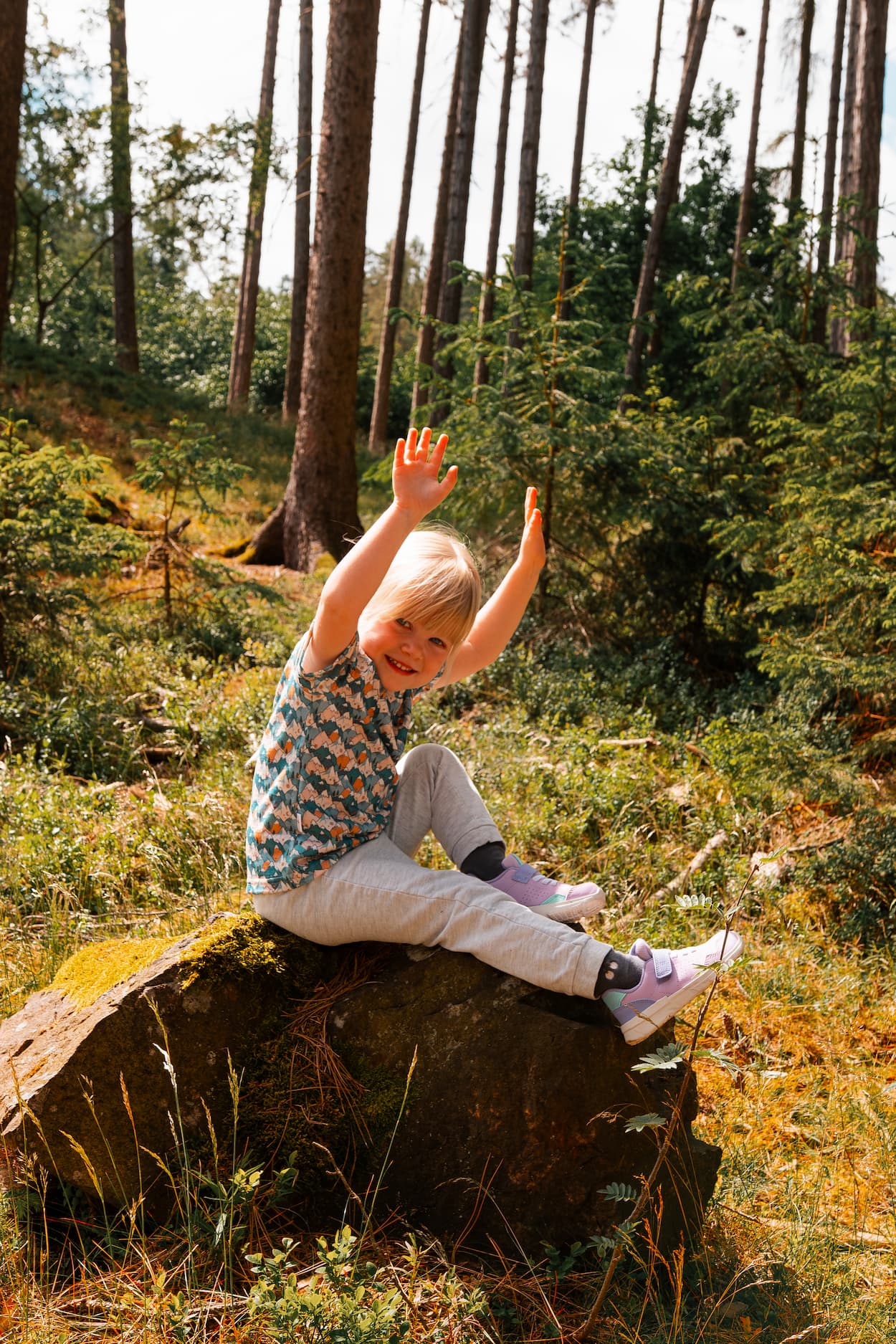Mädchen trägt ein Merino Kinder T-Shirt mit blauem Bergmotiv von Bellibell und sitzt auf einem Felsen im Wald.