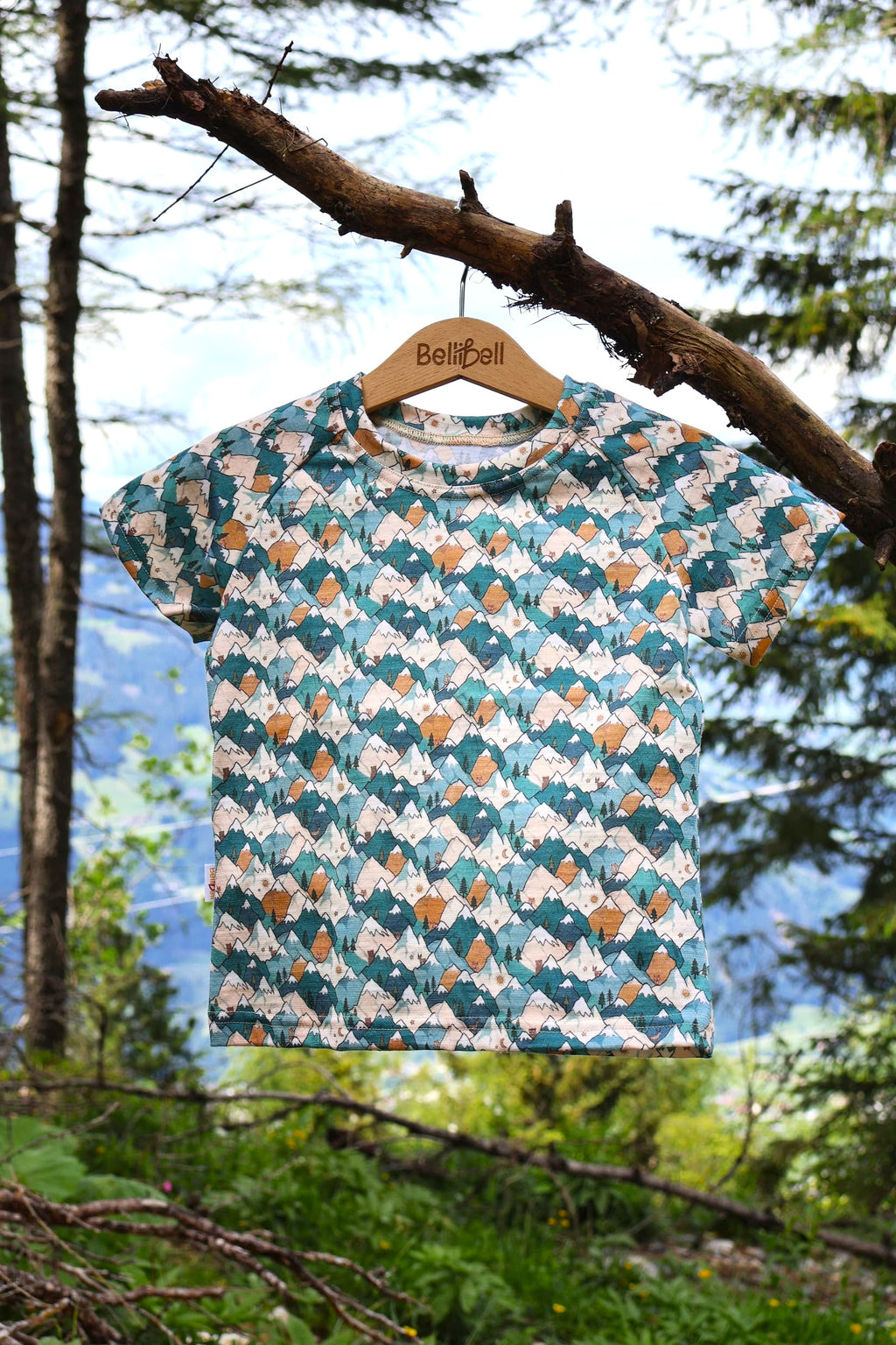 Merino Kinder-T-Shirt mit blauem Bergmotiv von Bellibell auf einem Kleiderbügel in den Bergen.